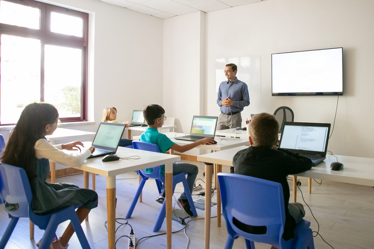 students at their computers in a classroom with a teacher at the front of the room